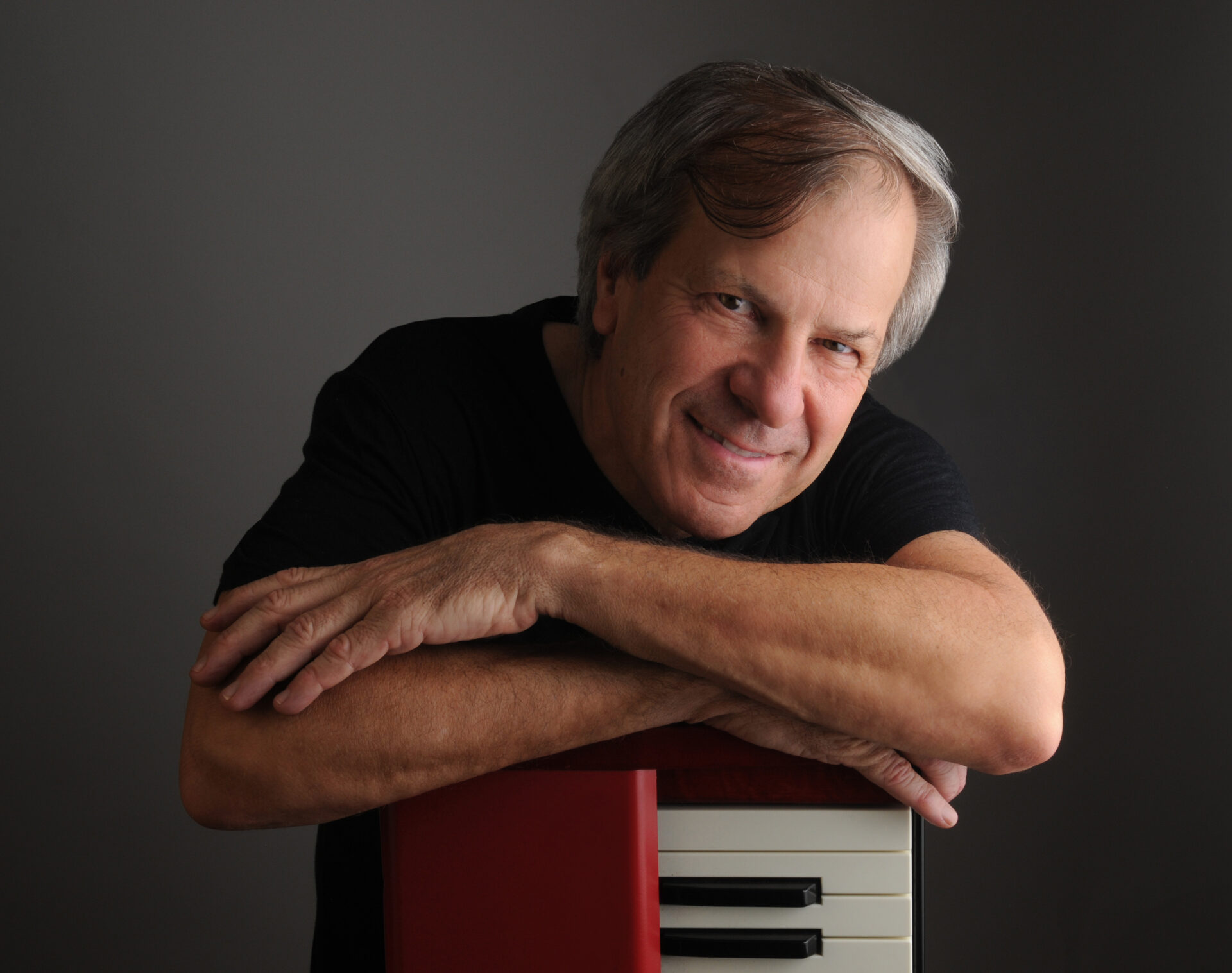 An older man with gray hair smiles warmly, leaning his arms on top of a red and white keyboard against a plain gray background. He wears a black shirt and looks directly at the camera.