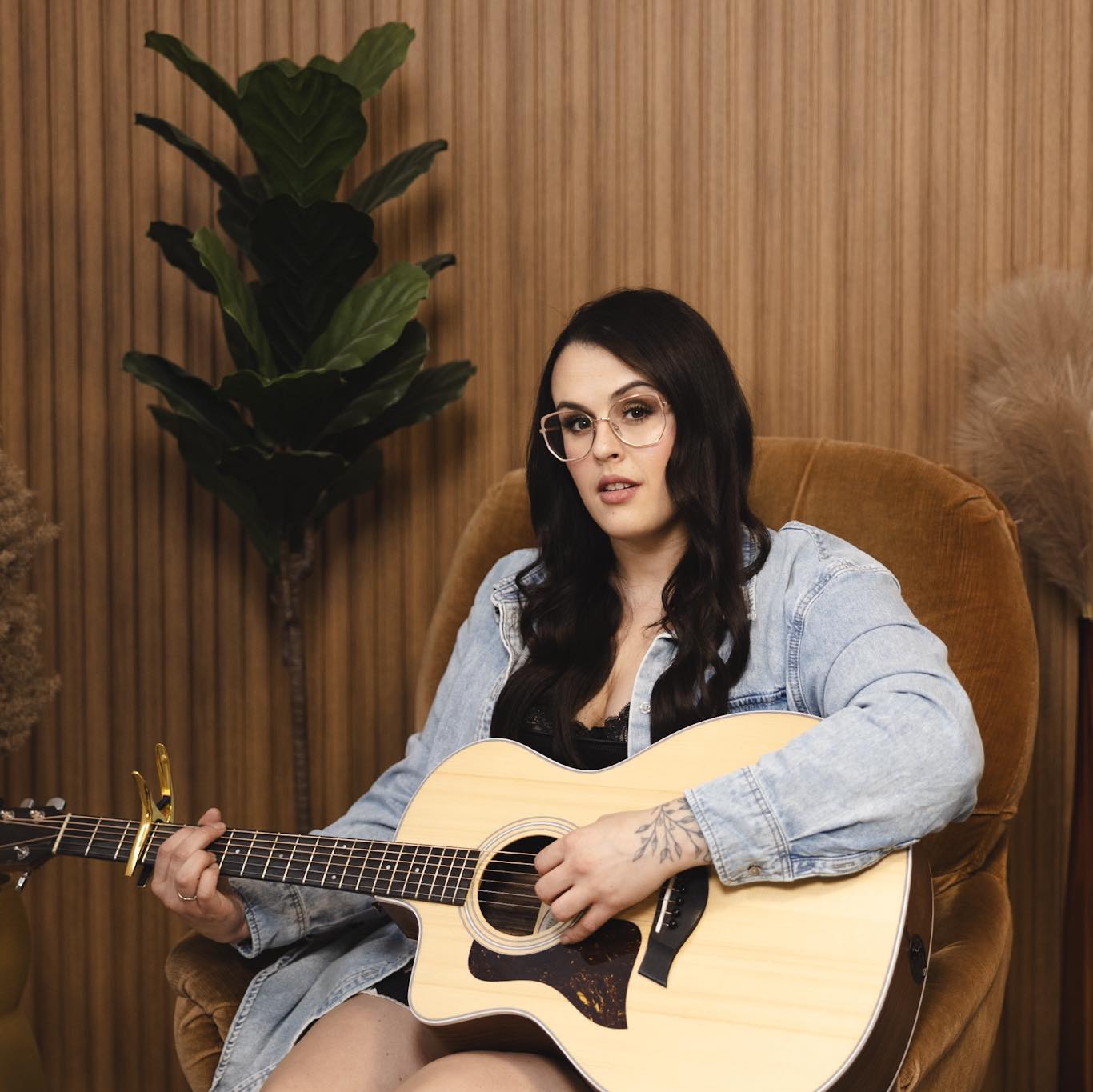 A woman with long dark hair and glasses sits on a brown chair, playing an acoustic guitar. She wears a denim jacket and is positioned in front of a wooden wall with a tall green plant nearby.