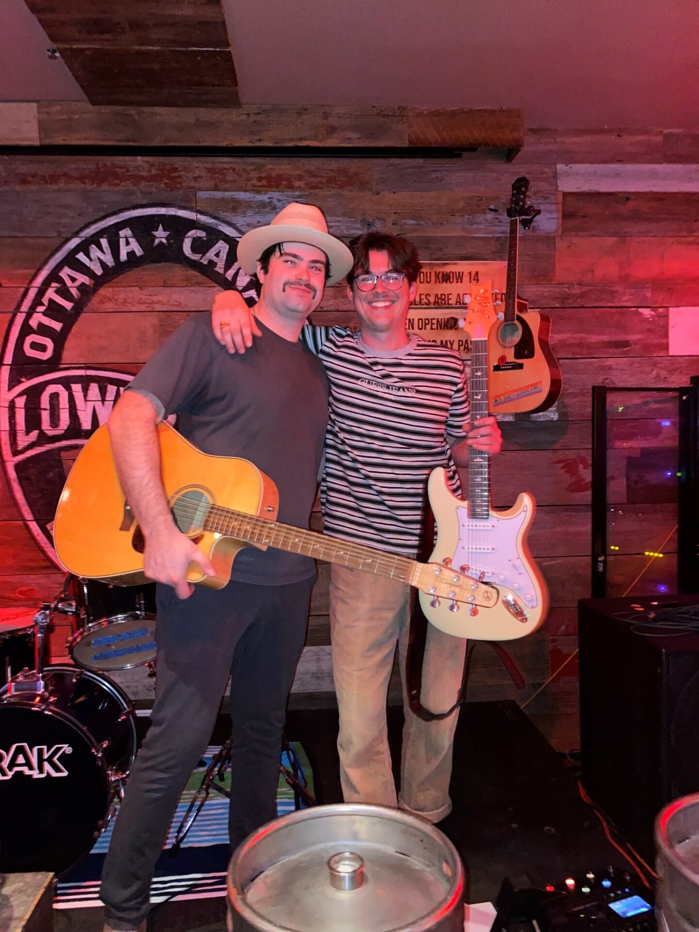 Two men stand smiling on a stage with their arms around each other, each holding an electric guitar. Drums and guitars are visible in the background, along with a wooden wall and an "Ottawa" sign.
