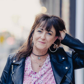 A woman with wavy brown hair wearing a black leather jacket, hoop earrings, and layered necklaces stands outdoors, smiling slightly and touching her hair. The background is softly blurred.