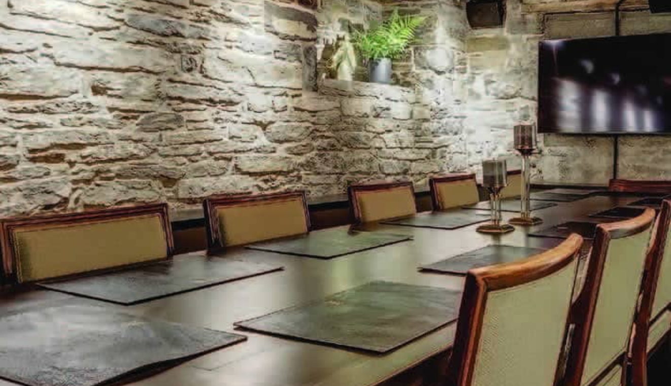 A long dining table with placemats and candles sits in a room with stone walls, cushioned chairs, a mounted TV, and a potted plant on a wall shelf, creating a cozy and rustic atmosphere.