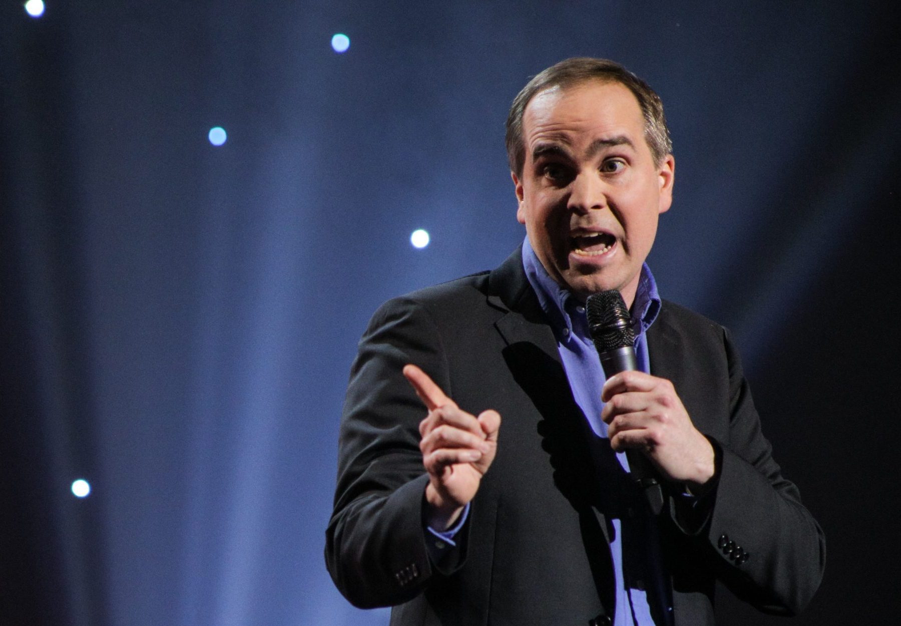 A man in a suit holds a microphone and gestures with his finger while speaking on stage, with bright spotlights and a dark background with small white lights behind him.