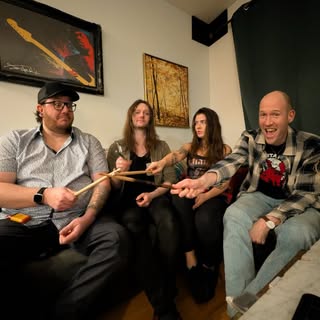 Four people sit on a couch, smiling and holding drumsticks together. Art hangs on the wall behind them. The group appears relaxed and happy, possibly band members posing for a casual photo.