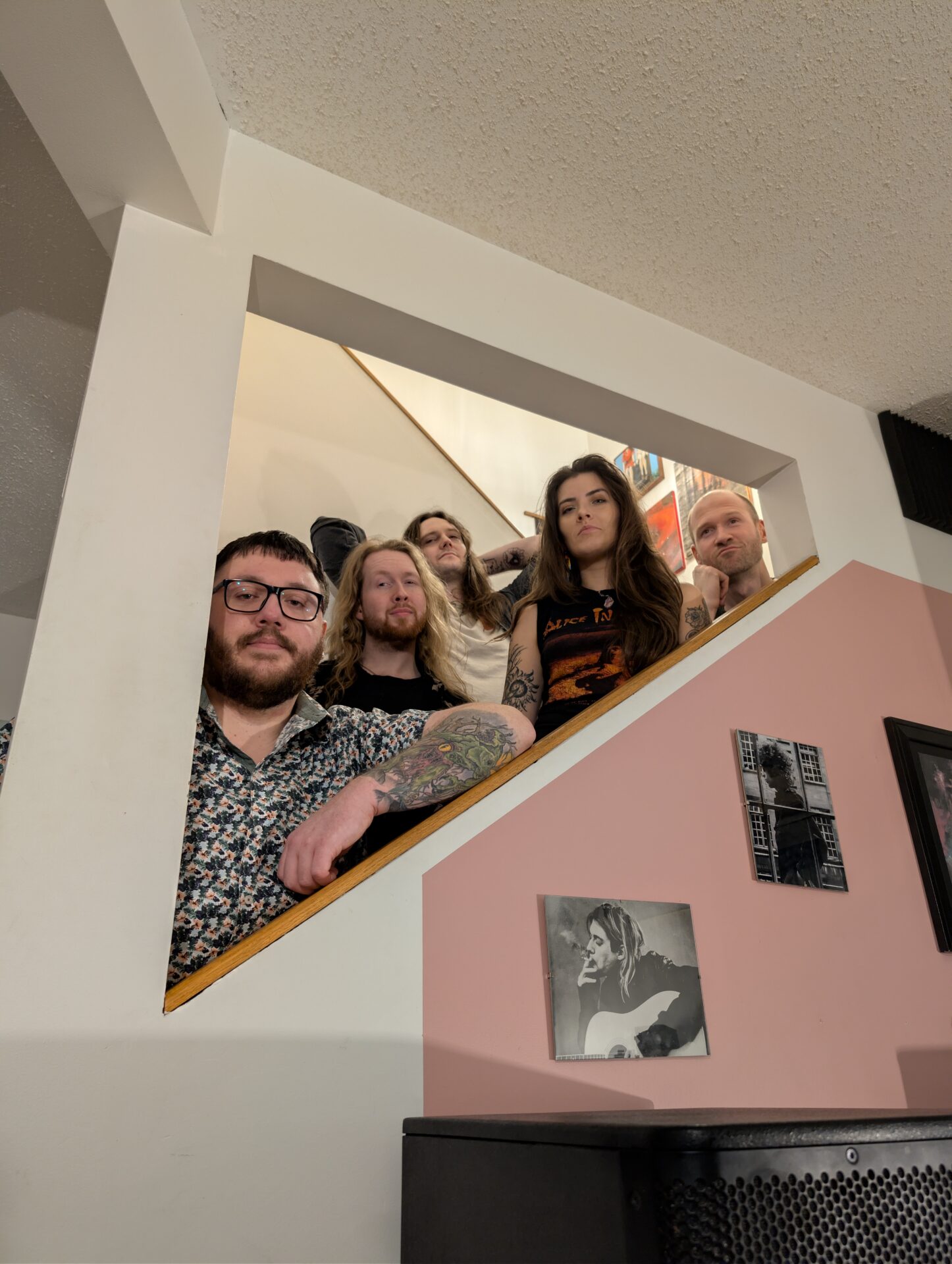 Five people stand on a staircase behind a triangular opening in a wall, looking toward the camera. Art and photos are visible on the walls in the foreground and background.