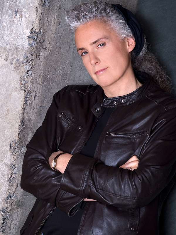 A person with short, curly gray hair, wearing a black leather jacket and a black headband, stands against a textured stone wall with arms crossed, giving a confident expression.