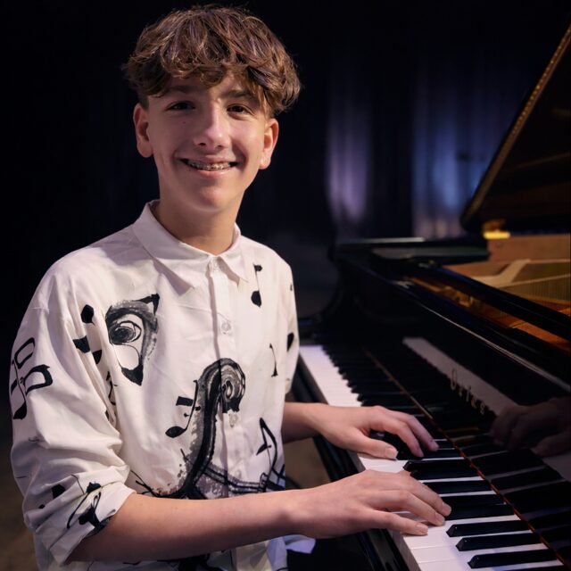 A smiling teenage boy with wavy brown hair plays a grand piano on stage. He wears a white shirt with artistic black designs and musical notes. The background is dark, highlighting the piano and the performer.
