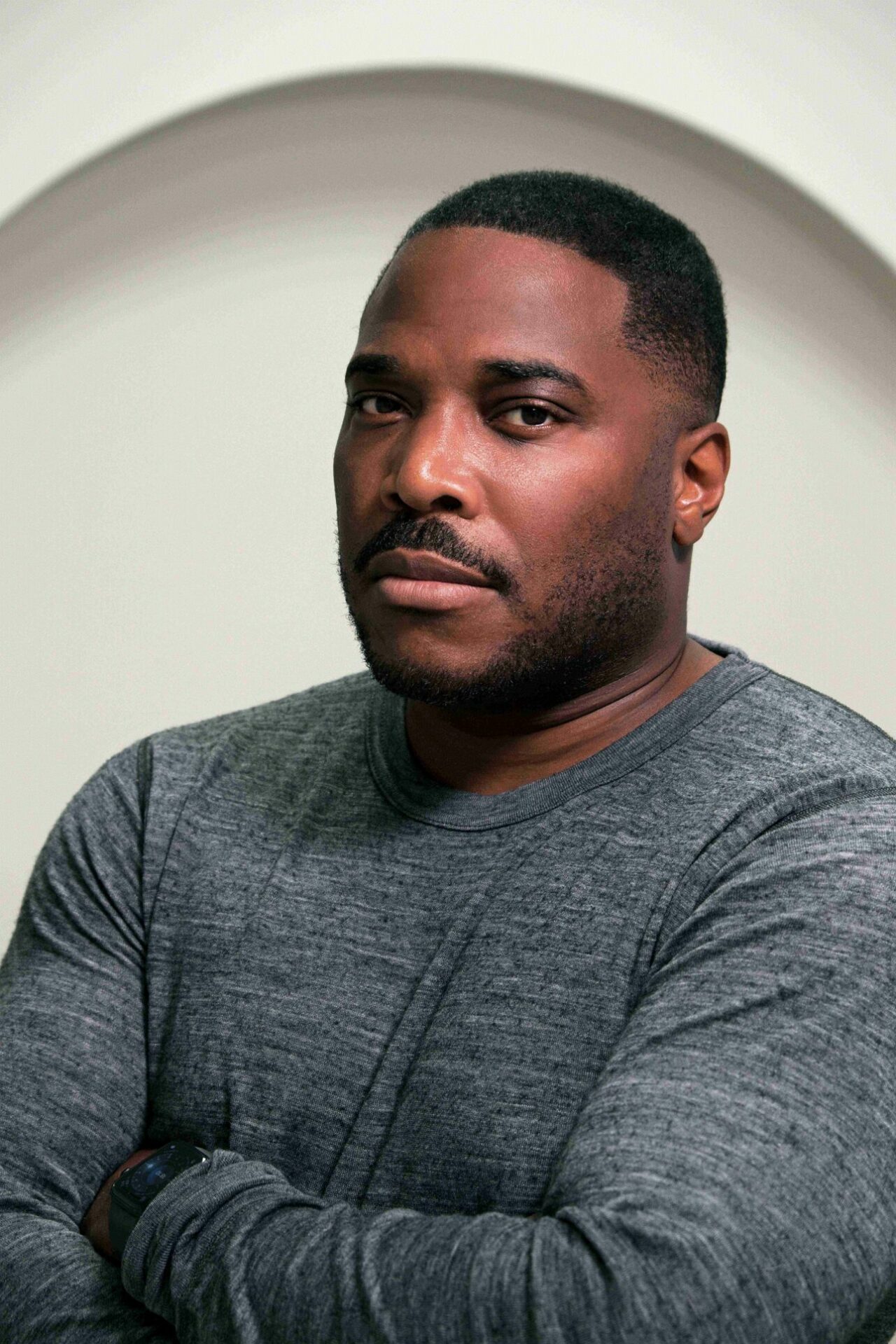 A man in a grey long-sleeve shirt stands with arms crossed, looking at the camera with a serious expression. The background is plain with a curved arch shape behind him.