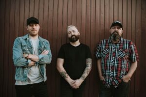 Three men stand in front of a vertical wooden wall. One wears a denim jacket and cap, one has a black shirt and visible tattoos, and the third wears a plaid shirt and trucker hat. All are looking at the camera.