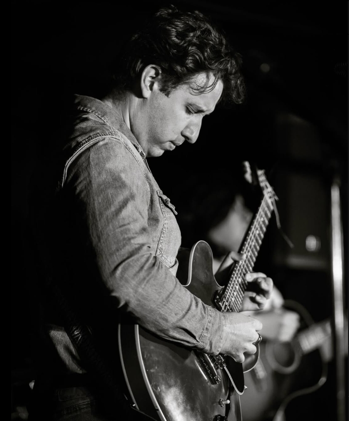 A man in a denim jacket plays an electric guitar with focus on stage. The image is in black and white, and another musician with a guitar is blurred in the background.