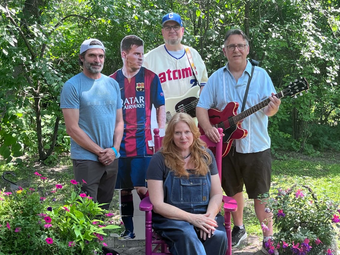 Four adults pose outdoors with a life-sized cardboard cutout of a soccer player; two men hold guitars, one woman sits in a pink chair, all surrounded by green trees and pink flowers.