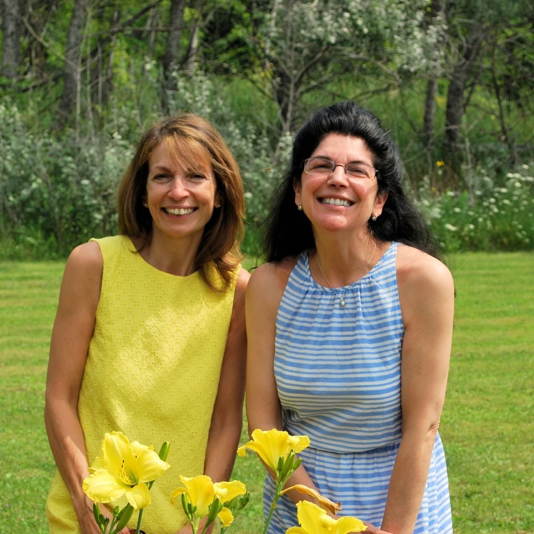 Two women smiling outdoors in a grassy area with trees in the background. One wears a yellow dress, the other wears a blue and white striped dress. Yellow flowers are in the foreground.